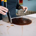 Close-up of a person tempering chocolate on a kitchen counter using a scraper and thermometer.